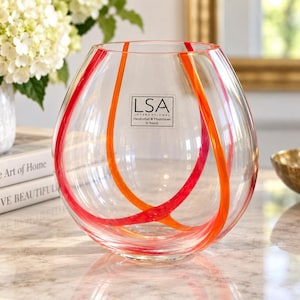 May include: A clear, spherical glass vase featuring bold, intersecting red and orange glass ribbons that wrap around the exterior. A square label on the front displays the text LSA INTERNATIONAL Handcrafted & Mouthblown in Poland.