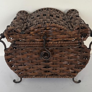 Puede incluir: Una cesta tejida marrón con tapa y asas de metal. La cesta tiene una forma curvada con un cierre y patas decorativas de metal. El patrón tejido es intrincado y la cesta parece estar hecha de materiales naturales.