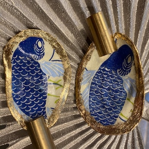 May include: Two decorative, oval-shaped objects with a blue fish design, outlined in gold leaf, and a brass tube. The fish has a blue scale pattern and is set against a cream and light blue background. The objects are set against a silver fan-like background.
