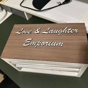 May include: A wooden box with a white label that says "Love & Laughter Emporium". The box has three drawers.