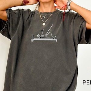 May include: A charcoal grey oversized t-shirt featuring a graphic of a skeleton performing pilates on a reformer machine. The shirt has short sleeves and a crew neckline. The word PEPPER is printed in black on the lower right side of the garment.