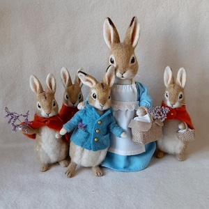 Puede incluir: Una familia de cinco figuritas de conejo de fieltro. La madre conejo lleva un vestido azul y un delantal blanco. Los otros conejos llevan chaquetas rojas y azules. Los conejos llevan cestas y flores.