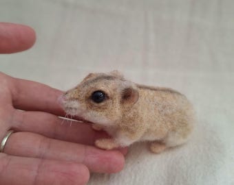 May include: A small, tan-colored felted hamster with dark eyes and small ears. The hamster is resting on a light-colored surface. The hamster is a handmade craft item.