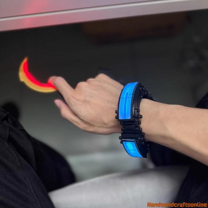 Puede incluir: Un reloj de pulsera futurista negro y azul con un dise&ntilde;o segmentado. El reloj presenta paneles iluminados de color azul brillante y una est&eacute;tica oscura e industrial. La imagen tambi&eacute;n muestra una forma de media luna roja brillante.