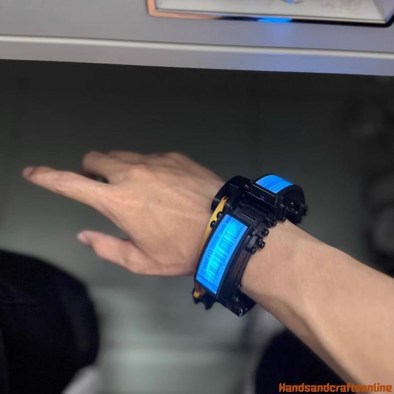 Puede incluir: Pulsera futurista negra y azul con una pantalla azul brillante. La pulsera tiene un dise&ntilde;o segmentado con un acento amarillo. La imagen est&aacute; tomada en interiores con un fondo neutro.