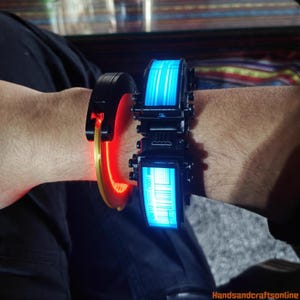 Puede incluir: Dos pulseras futuristas con elementos brillantes. Una es negra con un acento rojo y dorado, la otra es negra con una pantalla azul que muestra un c&oacute;digo de barras y l&iacute;neas verticales. Las pulseras se llevan en la mu&ntilde;eca.