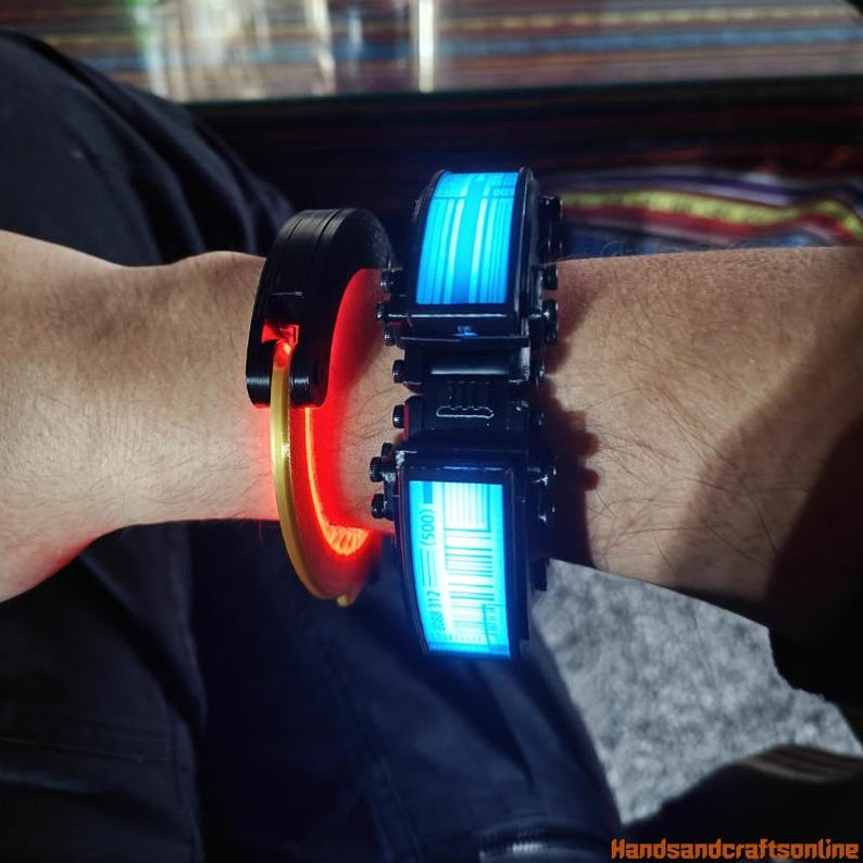 Puede incluir: Dos pulseras futuristas con elementos brillantes. Una es negra con un acento rojo y dorado, la otra es negra con una pantalla azul que muestra un c&oacute;digo de barras y l&iacute;neas verticales. Las pulseras se llevan en la mu&ntilde;eca.
