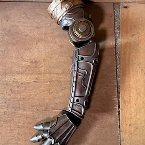 May include: A steampunk-inspired prosthetic arm in metallic tones of brown, bronze, and silver. The arm has articulated joints, a detailed forearm, and a hand with individual finger components. The design incorporates rivets and straps, reflecting a mechanical aesthetic.