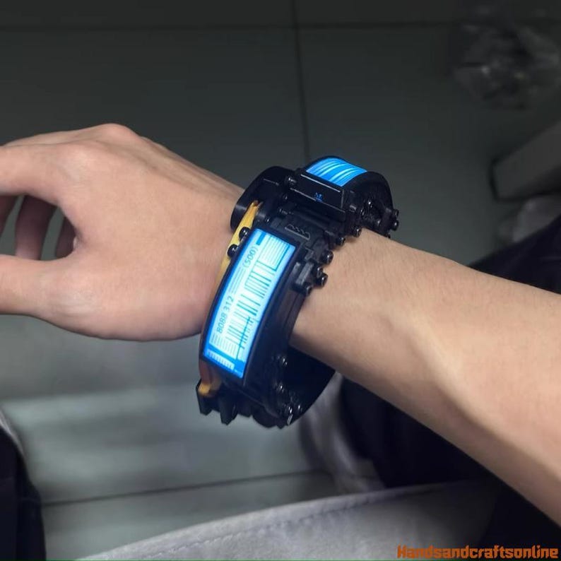 Puede incluir: Brazalete futurista negro y amarillo con una pantalla azul iluminada. El brazalete presenta un dise&ntilde;o de c&oacute;digo de barras y se lleva en la mu&ntilde;eca. El dise&ntilde;o recuerda a la tecnolog&iacute;a de ciencia ficci&oacute;n.