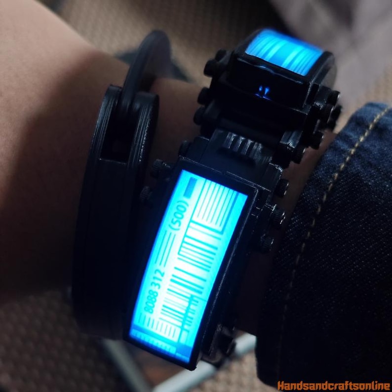 Puede incluir: Una pulsera futurista negra con dos pantallas rectangulares que muestran luz azul y patrones de c&oacute;digo de barras. La pulsera se asegura con un cierre negro. El texto "(500)" y "8088 312" son visibles en una pantalla. La pulsera se lleva en una mu&ntilde;eca.