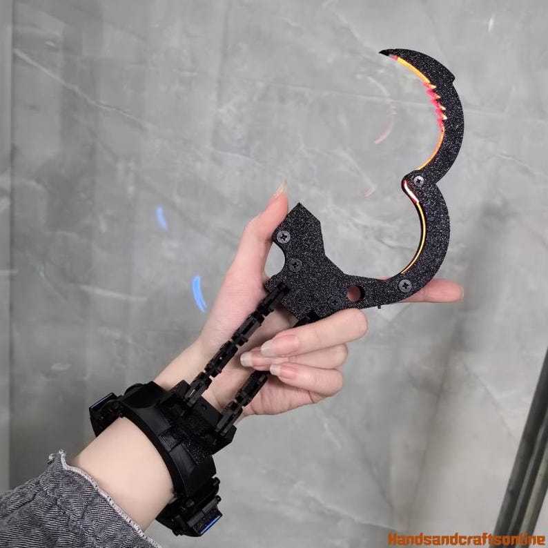 Puede incluir: Garra negra de mu&ntilde;eca con una cuchilla curva y un brazalete. La cuchilla tiene detalles en naranja y amarillo. La garra se sostiene en una mano sobre un fondo gris.