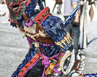 Könnte beinhalten: Ein detailliertes Cosplay-Kostüm eines Charakters aus der Monster Hunter-Videospielserie. Das Kostüm verfügt über einen Helm mit blauen und roten Federn, eine blaue und violette Rüstung mit einem braunen Ledergürtel und eine große, detaillierte Waffe.