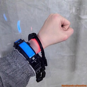 Puede incluir: Un dispositivo de mu&ntilde;eca futurista negro y azul con una pantalla azul rectangular y una luz roja. El dispositivo se lleva en la mu&ntilde;eca de una persona, con una correa negra y detalles mec&aacute;nicos intrincados.