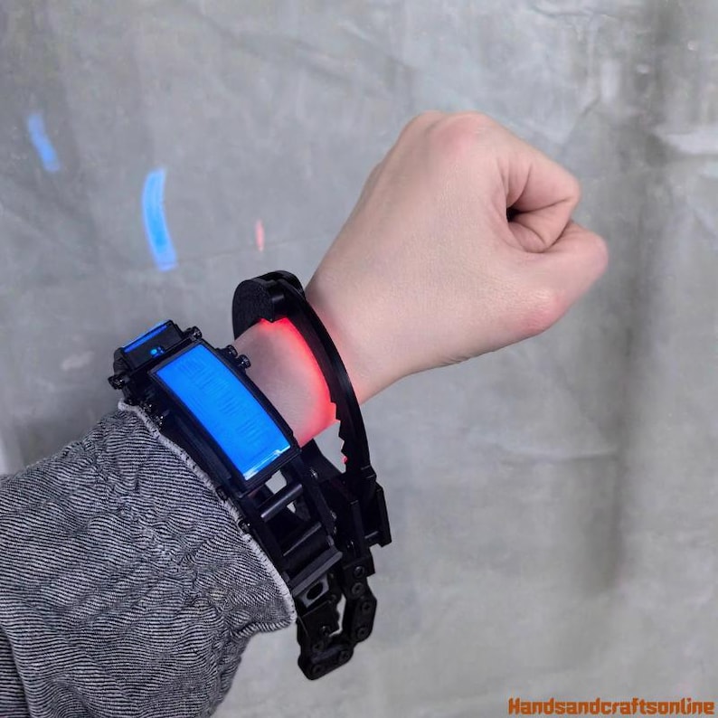 Puede incluir: Un dispositivo de mu&ntilde;eca futurista negro y azul con una pantalla azul rectangular y una luz roja. El dispositivo se lleva en la mu&ntilde;eca de una persona, con una correa negra y detalles mec&aacute;nicos intrincados.