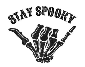 May include: Black and white graphic design of a skeleton hand giving the shaka sign with the text "STAY SPOOKY" above it.