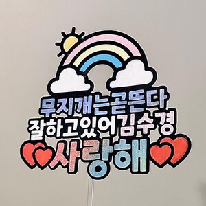 May include: A colorful cake topper featuring a rainbow, sun, and clouds. The topper has Korean text and two red hearts. The text includes the words "Love" and "Kim Soo Kyung".