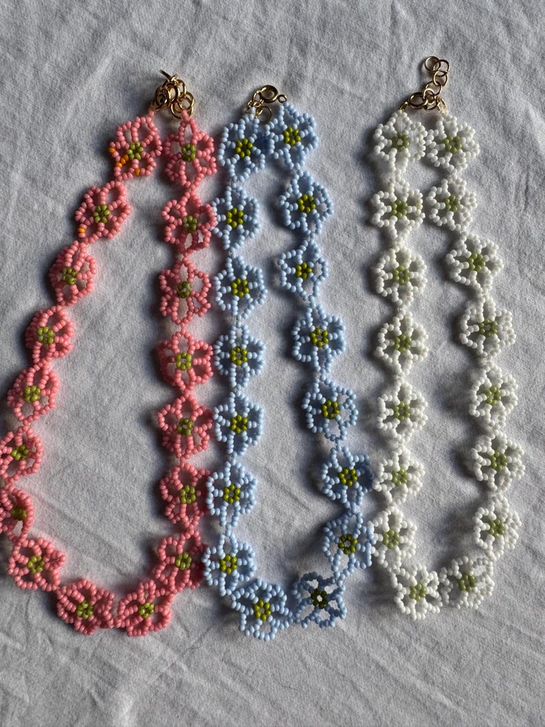 Op de afbeelding: Drie kralenkettingen in roze, lichtblauw en wit, elk met een bloemmotief. De bloemen hebben groene centra en zijn aan elkaar geregen met goudkleurige sluitingen. De kettingen worden op een wit oppervlak gepresenteerd.