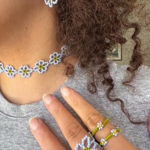 Op de afbeelding: Een kralen sieradenset met een ketting, oorbellen en ringen. De sieraden hebben een madeliefbloemdesign met lichtblauwe, gele en groene kralen. De oorbellen hangen aan goudkleurige haken.