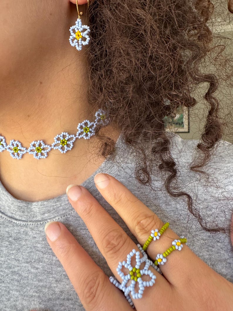 Op de afbeelding: Een kralen sieradenset met een ketting, oorbellen en ringen. De sieraden hebben een madeliefbloemdesign met lichtblauwe, gele en groene kralen. De oorbellen hangen aan goudkleurige haken.