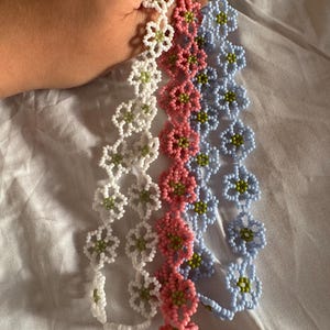 Op de afbeelding: Vier bloemenarmbanden van kralen in wit, roze en lichtblauw. Elke armband heeft een bloemenontwerp met groene centra. De armbanden worden tegen een witte achtergrond gehouden.