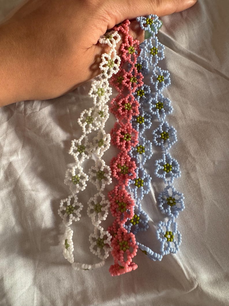 Op de afbeelding: Vier bloemenarmbanden van kralen in wit, roze en lichtblauw. Elke armband heeft een bloemenontwerp met groene centra. De armbanden worden tegen een witte achtergrond gehouden.