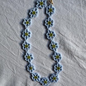 Op de afbeelding: Een ketting van lichtblauwe bloemvormige kralen met gele centra. De ketting heeft een goudkleurige sluiting en wordt gepresenteerd op een witte ondergrond. Het bloemendesign voegt een delicate touch toe aan deze sieraden.