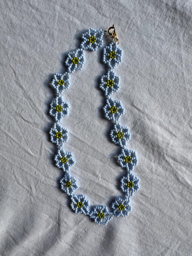 Op de afbeelding: Een ketting van lichtblauwe bloemvormige kralen met gele centra. De ketting heeft een goudkleurige sluiting en wordt gepresenteerd op een witte ondergrond. Het bloemendesign voegt een delicate touch toe aan deze sieraden.