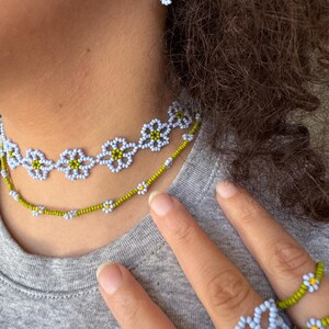 Op de afbeelding: Een kralen sieradenset met een ketting, oorbellen en ringen. De sieraden zijn gemaakt met lichtblauwe, witte en gele kralen, gerangschikt in een madeliefpatroon. De ketting en ringen zijn zichtbaar in de afbeelding.
