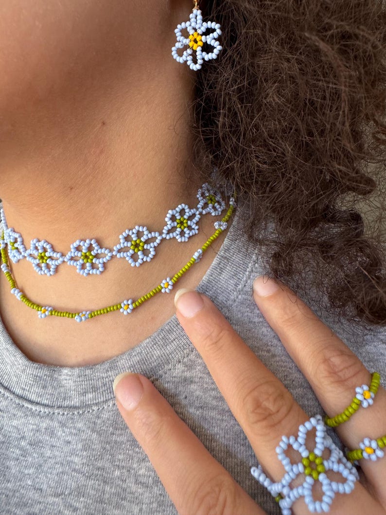 Op de afbeelding: Een kralen sieradenset met een ketting, oorbellen en ringen. De sieraden zijn gemaakt met lichtblauwe, witte en gele kralen, gerangschikt in een madeliefpatroon. De ketting en ringen zijn zichtbaar in de afbeelding.