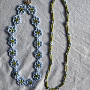 Op de afbeelding: Twee kralenkettingen. E&eacute;n ketting heeft lichtblauwe bloemvormige kralen met gele centra. De andere ketting is een streng kleine witte en gele bloemkralen op een groene draad. Beide kettingen hebben gouden sluitingen.