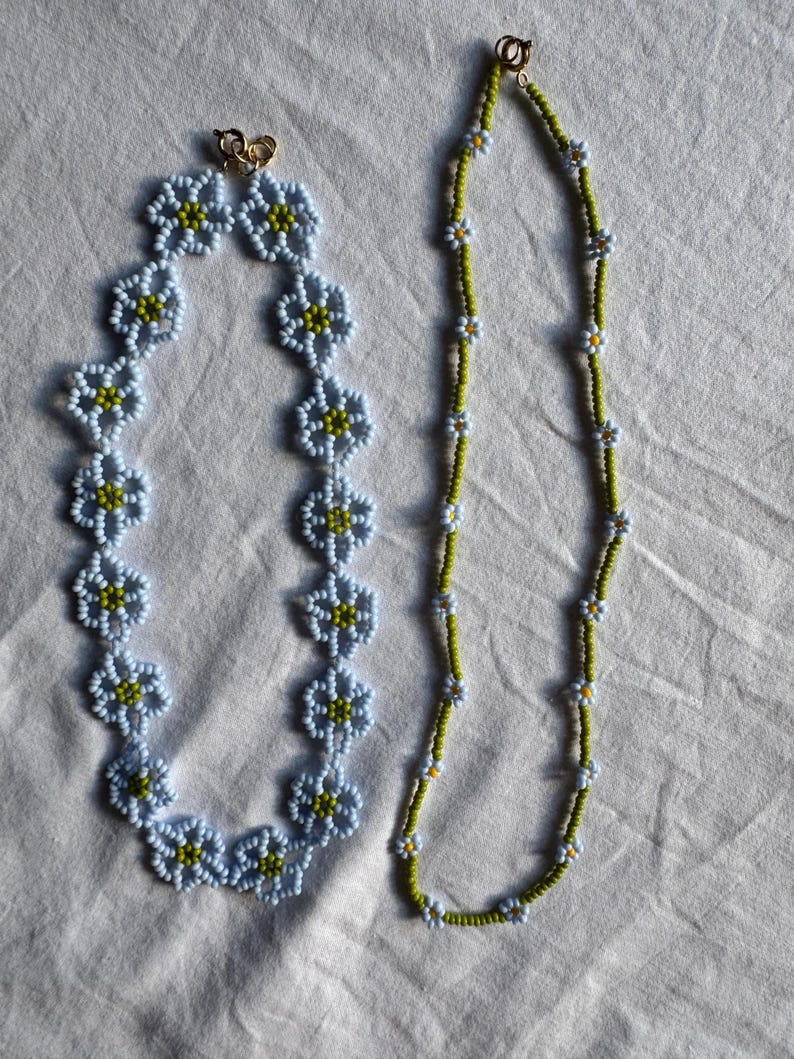 Op de afbeelding: Twee kralenkettingen. E&eacute;n ketting heeft lichtblauwe bloemvormige kralen met gele centra. De andere ketting is een streng kleine witte en gele bloemkralen op een groene draad. Beide kettingen hebben gouden sluitingen.