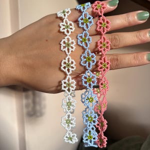 Op de afbeelding: Drie kralenarmbanden in wit, lichtblauw en roze, elk met een bloemenmotief. De bloemen hebben groene centra. De armbanden worden tegen een neutrale achtergrond gepresenteerd.