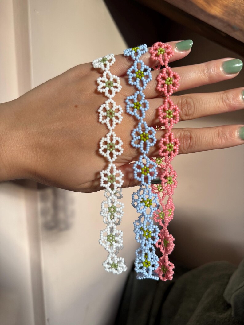 Op de afbeelding: Drie kralenarmbanden in wit, lichtblauw en roze, elk met een bloemenmotief. De bloemen hebben groene centra. De armbanden worden tegen een neutrale achtergrond gepresenteerd.