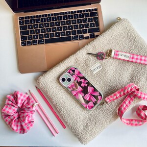 Könnte beinhalten: Ein roségoldenes Laptop liegt neben einer beigefarbenen, flauschigen Tasche, einer Handyhülle mit rosa Schleifen und einem rosa-weiß karierten Haargummi. Ein rosa Schlüsselband und Bleistifte sind ebenfalls zu sehen. Das Wort "lümina" ist aufgedruckt.