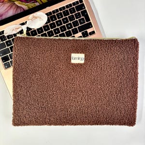 Peut inclure: Une housse d'ordinateur portable marron texturée avec une fermeture éclair dorée et une petite étiquette "lúmina". Le manchon est posé sur un ordinateur portable rose doré.