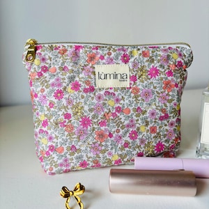 Könnte beinhalten: Eine Kosmetiktasche mit floralem Muster, einem goldenen Reißverschluss und dem Markennamen "lúmina" auf einem Etikett. Die Tasche hat ein Muster aus rosa, orangefarbenen und gelben Blumen auf weißem Grund. Eine goldene Schleife und Make-up-Artikel sind im Vordergrund.