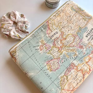 Könnte beinhalten: Eine Reißverschluss-Reisetasche mit Vintage-Kartendesign, goldenem Reißverschluss und einem Stoffetikett mit der Aufschrift "lumina". Ein floraler Scrunchie und eine kleine Dose sind ebenfalls zu sehen. Die Tasche ist ca. 30 cm lang.