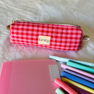 Può includere: Un astuccio a quadretti rossi e rosa con cerniera dorata e una piccola etichetta con la scritta "lúmina lovers". Nell'immagine sono presenti anche una cartella rosa e pennarelli colorati.