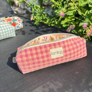 Può includere: Un astuccio a quadretti vichy rosa e arancione chiaro con chiusura a zip e un'etichetta in tessuto beige con la scritta "lùmina lovers" in nero. La fodera interna presenta un motivo floreale con disegni botanici rosa, arancioni e gialli.