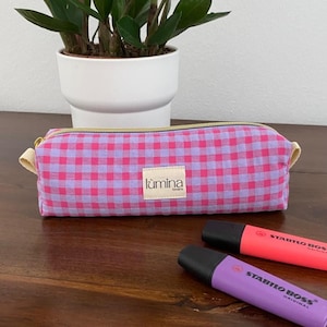 Può includere: Astuccio a quadretti rosa e viola con cerniera dorata e passanti in tessuto color crema. L'astuccio rettangolare ha una piccola etichetta bianca con la scritta "lùmina". Due evidenziatori, uno rosa e uno viola, sono accanto.