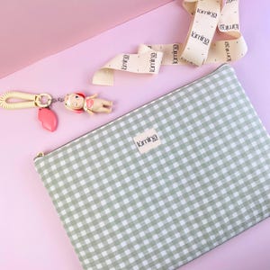 Puede incluir: Un estuche con cremallera de cuadros verdes y blancos con una pequeña etiqueta que dice "lúmina". Un llavero con un dije de corazón rosa y una muñeca pequeña está al lado del estuche. Una cinta con la palabra "lúmina" también está en la imagen.