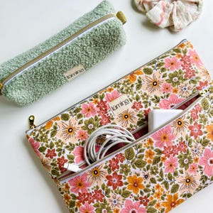 Peut inclure: Une trousse verte et duveteuse et une pochette zippée à imprimé floral avec un chargeur et un câble blancs à l'intérieur. La pochette florale présente un motif de fleurs roses, oranges et vertes. Les deux articles ont des fermetures éclair dorées et la marque "lūmina".