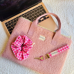 Può includere: Una borsa rosa per laptop con un elastico per capelli e un portachiavi abbinati. La borsa ha un esterno strutturato e sfocato, una chiusura con cerniera e un manico a spina di pesce. L'elastico e il braccialetto presentano un motivo a quadri rosa e bianco. Un laptop dorato è sullo sfondo.