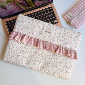 Peut inclure: Une housse d'ordinateur portable à motifs floraux avec une volants en vichy rose et une trousse assortie. La housse et l'étui ont un fond crème avec de petites fleurs roses. Le nom de la marque "lúmina" est sur une petite étiquette.