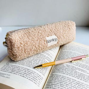 Puede incluir: Un estuche beige y esponjoso con cremallera dorada y una pequeña etiqueta blanca que dice "lumina lovers". El estuche descansa sobre un libro abierto junto a un bolígrafo con detalles amarillos, rosas y dorados.