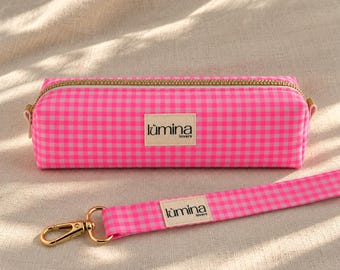 Puede incluir: Un estuche rectangular con un patrón de cuadros vichy en rosa brillante y blanco, con una cremallera dorada y una correa de muñeca a juego con un mosquetón dorado. Una etiqueta de tela beige en el estuche y la correa dice lùmina lovers.