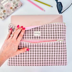 Könnte beinhalten: Eine rechteckige Laptoptasche mit einem braun-hellrosa Vichy-Karo-Muster. Die Tasche verfügt über einen Reißverschluss an der Oberseite und eine vordere Reißverschlusstasche mit hellrosa Innenfutter. Ein weißes Etikett mit dem Text lumina lovers ist mittig.