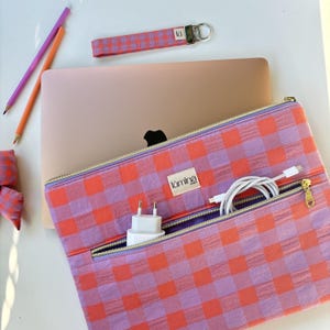 Peut inclure: Une housse pour ordinateur portable à motif vichy rose et orange, dotée d'une poche frontale zippée contenant un adaptateur secteur blanc et un câble de charge blanc enroulé. Un porte-clés en tissu assorti et deux crayons sont posés à côté.