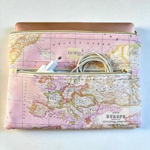 Puede incluir: Una funda rosa para portátil con diseño de mapa, cremalleras doradas y el logo "lümina". La funda tiene un bolsillo frontal que contiene un cargador blanco y un cable enrollado. Un portátil es parcialmente visible.