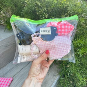 Peut inclure: Une pochette transparente en plastique avec une fermeture éclair et un bord vert néon contenant des lunettes de soleil sombres, un chouchou à carreaux vichy rose et blanc, un masque en tissu rose clair texturé avec l'étiquette Lumina Lovers.
